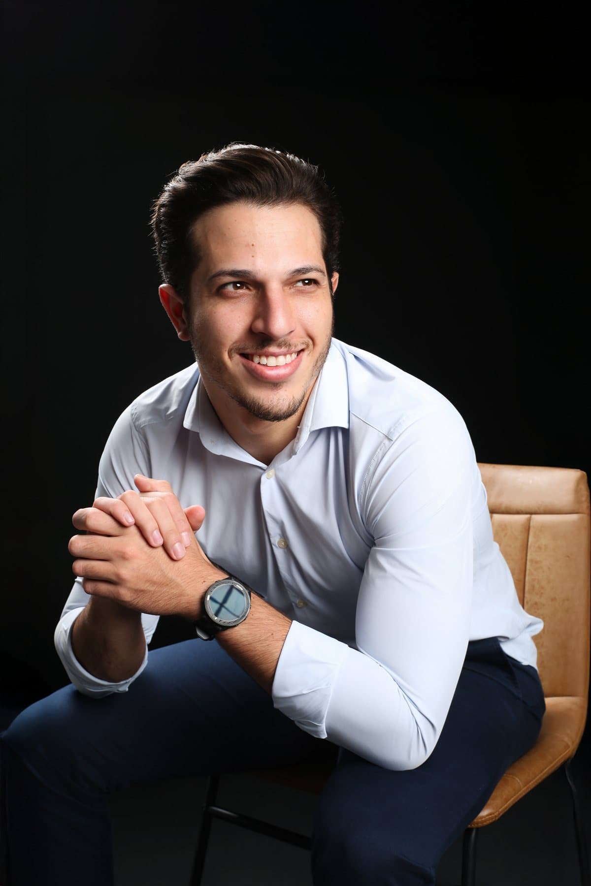 Dr. Guilherme Magela sentado, sorrindo, de camisa social azul clara e mãos entrelaçadas, em retrato de estúdio
