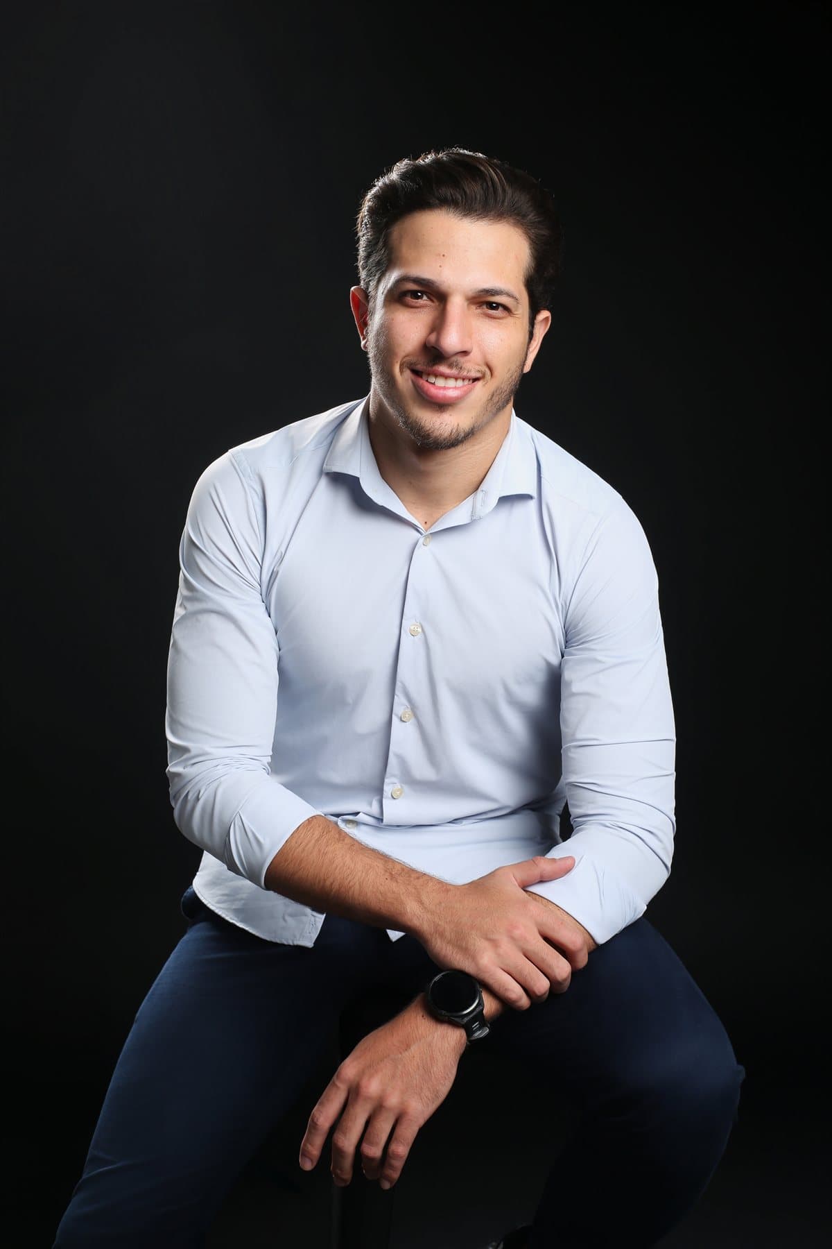 Dr. Guilherme Magela sentado, sorrindo, de camisa social azul clara e braços cruzados, em retrato de estúdio